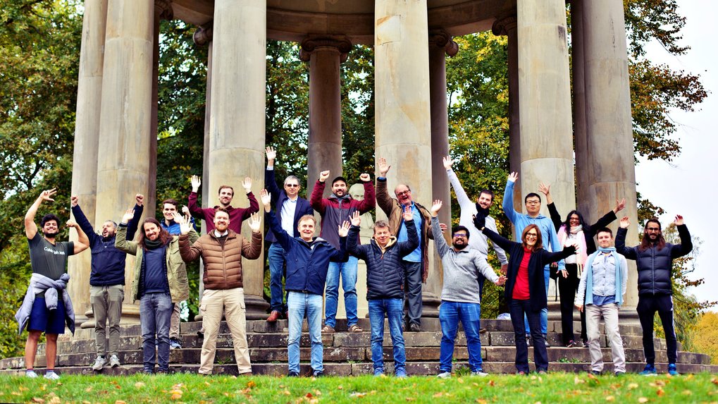 Eine der Kamera zugewandte Gruppe von Personen steht vor einem tempelartigen Rundbau mit grauen Steinsäulen. Alle Personen haben ihre Hände erhoben und winken.