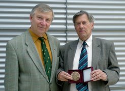 Jiří Bičák and Jürgen Ehlers stand with a medal in front of a gray background.